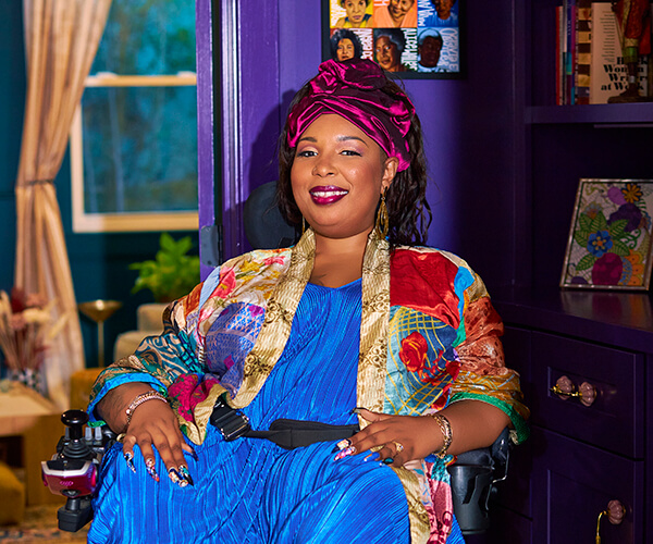 Andraéa LaVant, a pecan-brown Black woman, smiles wide at the camera from the side while seated at a desk. She wears a bright blue jumper and a colorful patchwork silk kimono, with loose braids wrapped in a deep fuchsia headwrap. Behind her is a purple office with books, a painting, a window, and curtains.