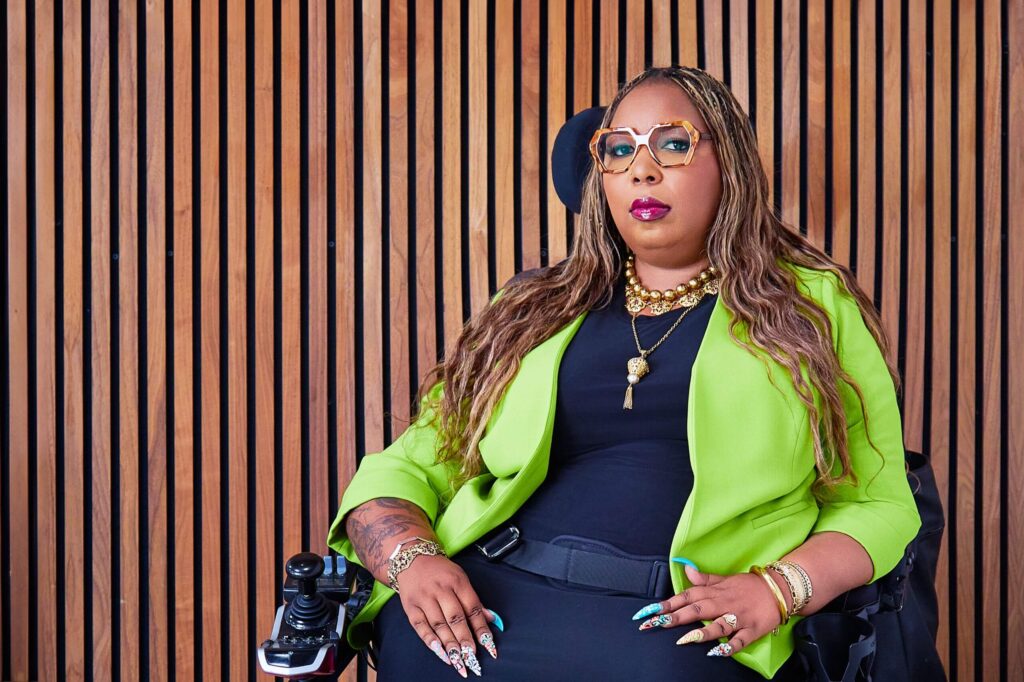Andraéa LaVant, a pecan-brown Black woman, looks smug and powerful, seated in her wheelchair with the headrest and joystick visible. Her ombré-orange hexagon glasses and fuchsia gloss complement wavy, two-tone mermaid braids cascading down her arms. A chartreuse blazer, black dress, jeweled nails, and gold jewelry stand out against a brown slatted wood backdrop.