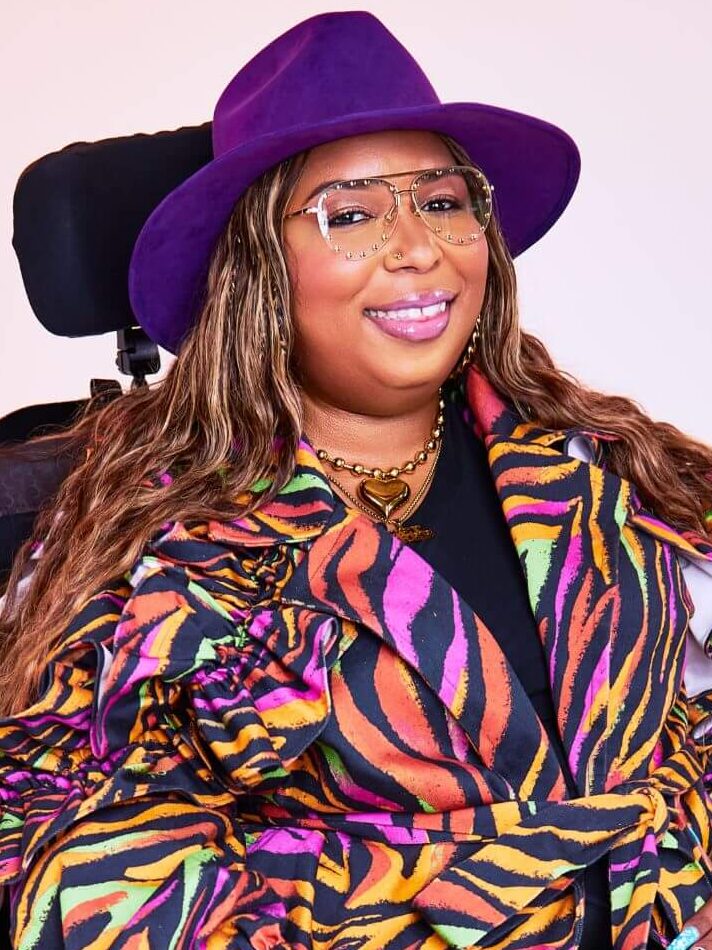 Andraéa LaVant, a pecan-brown Black woman, smiles wide in pink lip gloss, a royal purple fedora, and studded aviator glasses. A belted neon zebra trench, gold beaded necklace with a heart pendant, and a hint of black dress stand out against a soft pink-purple backdrop, with her headrest peeking through.