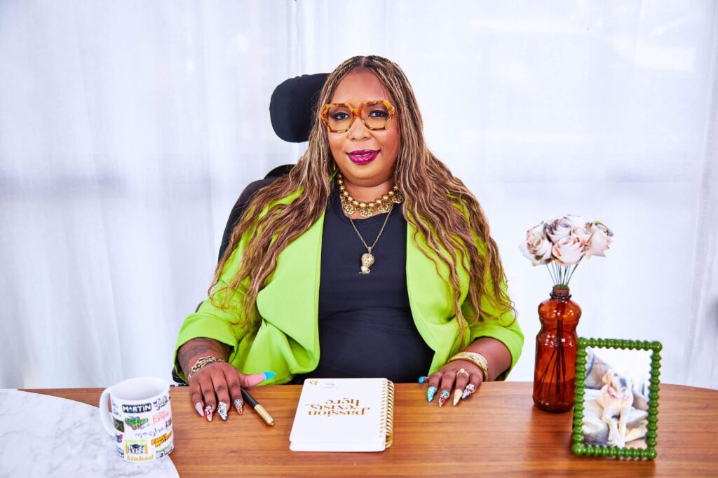 Andraéa LaVant, a pecan-brown Black woman in ombré-orange glasses, gold jewelry, jeweled nails, and a chartreuse blazer, sits behind an executive desk slightly smiling. Her mermaid braids fall over her shoulders. In front of her are a mug with 90s TV logos, a journal, paper flowers in a bottle vase, and a framed photo of her dog, Goji, with her wheelchair subtly visible.